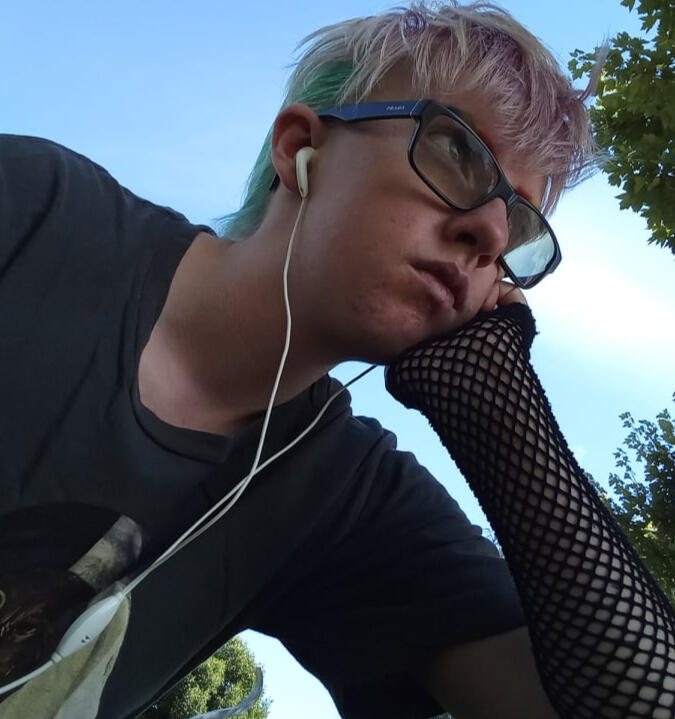 An image of a trans guy with pink and blue hair and earphones in with a background of a blue sky and a tree