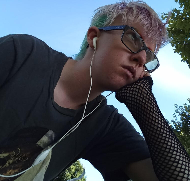 An image of a trans guy with pink and blue hair and earphones in with a background of a blue sky and a tree